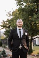 retrato de el novio en naturaleza en el verano. elegante alto novio en un elegante negro negocio traje. empresario en naturaleza. retrato de un exitoso hombre. Boda retrato de el novio. foto