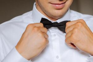 A bearded man in a white shirt adjusts his bow tie. Groom's morning. Close-up detail, men's tie for a wedding or an important meeting. photo