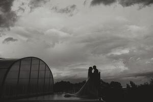 Silhouettes of the bride and groom in full height, standing against the background of a voluminous cloudy sky. Black and white photo. photo