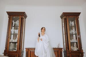 un hermosa joven novia niña sostiene un elegante botella de perfume en su mano y es Listo a rociar sí misma. Mañana de el novia antes de el Boda ceremonia. foto