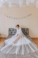 Beautiful bride in white wedding dress sitting on the bed in the morning. Fashion shot, beautiful woman in a dressing gown. Fashion, glamor concept photo
