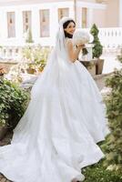 Photo from the back. A beautiful young woman in a white wedding dress is smiling on a warm wedding summer day