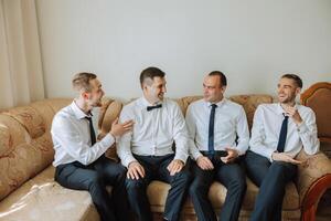 elegante novio riendo y teniendo divertido con padrinos de boda mientras consiguiendo Listo en el Mañana para Boda ceremonia. lujo hombre foto