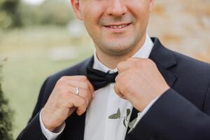 Portrait of the groom in nature in the summer. A stylish man in an elegant black business suit is tying a bow tie, close-up photo. Businessman. Portrait of a successful man. Wedding portrait. photo