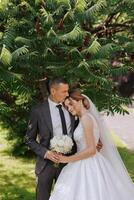 A handsome groom embraces his bride in a lush white dress and smiles in a beautiful outdoor setting. Under the open sky. High quality photo. A newlywed couple poses together on a sunny summer day. photo