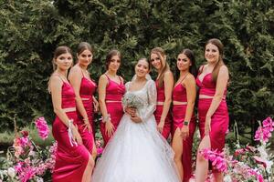 A brunette bride and her bridesmaids in matching pink dresses are standing near the solemn arch. Girls in identical dresses are making out at a wedding. Wedding in nature. photo
