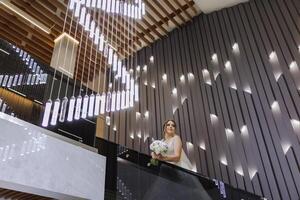 un hermosa novia en un vestir con un largo tren es en pie en el escaleras. blanco Boda vestido. hermosa interior. foto