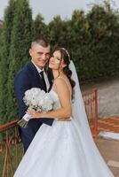 A handsome groom embraces his bride in a lush white dress and smiles in a beautiful outdoor setting. Under the open sky. High quality photo. A newlywed couple poses together on a sunny summer day. photo