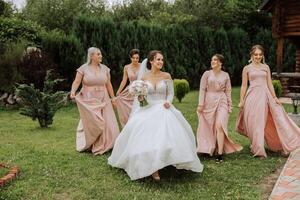 A brunette bride and her bridesmaids in identical pink dresses are walking against the background of nature. Girls in identical dresses are making out at a wedding. Wedding in nature. photo