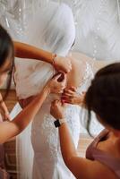 Wedding morning. Bridesmaids help put on the white wedding dress. A young woman is preparing to meet her groom and having fun with her friends photo