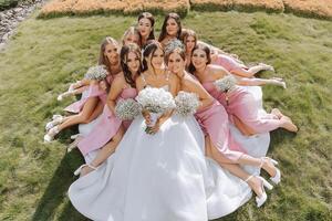 A brunette bride and her bridesmaids in identical dresses sit with the bride and rejoice with flowers in their hands on the green grass. Wedding in nature. photo