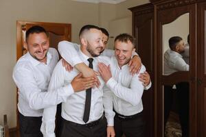 elegante novio riendo y teniendo divertido con padrinos de boda mientras consiguiendo Listo en el Mañana para Boda ceremonia. lujo hombre foto