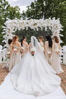 The bride and her bridesmaids turned their backs to the camera against the background of green trees. A long train on the dress. The girls are wearing identical dresses at the wedding. photo