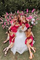 un morena novia y su damas de honor en vestidos de el mismo color sentar con el novia y alegrarse con flores en su manos cerca el solemne arco. Boda en naturaleza. foto