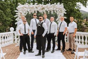 alegre, joven, energético testigos de el novio siguiente a el novio. amigos felicitar el novio. el novio en un chaleco y su amigos en blanco camisas actitud cerca el Boda arco. Boda en naturaleza. foto