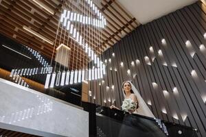 A beautiful bride in a dress with a long train is standing on the stairs. White wedding dress. Beautiful interior. photo