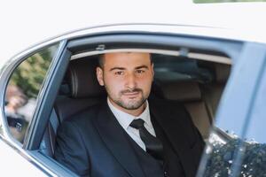 a handsome groom sits with a wedding bouquet inside a white car and looks out the window. A happy moment on the wedding day. photo