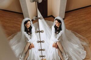 un hermosa novia es sentado en un vendaje vestido en el Mañana antes de el Boda ceremonia en un hotel con un moderno interior. increíble peinado de el novia. natural y moderno constituir. foto