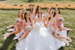 A brunette bride and her bridesmaids in identical dresses sit with the bride and rejoice with flowers in their hands on the green grass. Wedding in nature. photo