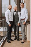 elegante novio riendo y teniendo divertido con padrinos de boda mientras consiguiendo Listo en el Mañana para Boda ceremonia. lujo hombre foto