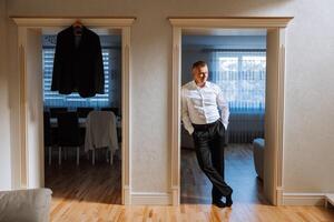 retrato de un joven novio a hogar antes de el Boda ceremonia. un hermoso hombre en un blanco camisa y negro pantalones es en pie en su habitación. foto