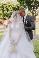 A handsome groom embraces his bride in a lush white dress and smiles in a beautiful outdoor setting. Under the open sky. High quality photo. A newlywed couple poses together on a sunny summer day. photo