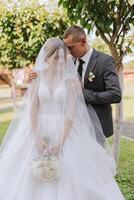 A handsome groom embraces his bride in a lush white dress and smiles in a beautiful outdoor setting. Under the open sky. High quality photo. A newlywed couple poses together on a sunny summer day. photo