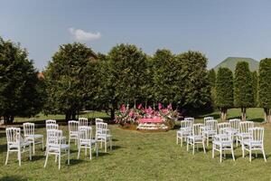 The pink stage is decorated with flowers on the background of the forest. Many white chairs on green grass. Preparation for the wedding ceremony photo