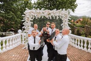 Cheerful, young, energetic witnesses of the bridegroom hold the bridegroom in their arms. Funny photo. The groom in a vest and his friends in white shirts pose near the wedding arch. Wedding in nature photo