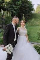 A handsome groom and an elegant bride in a lush white dress are walking in a summer park. Happy bride and groom getting ready for their best day. photo