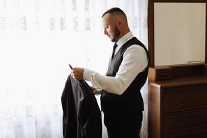 Portrait of a young groom with a beard at home before the wedding ceremony. Handsome man with a black beard, wearing a classic suit. Male portrait. photo