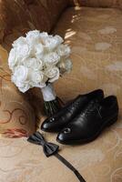 On the floor are men's dark leather shoes and a black belt, a man's suit on a mannequin, a wedding bouquet of flowers, wedding rings, men's perfume. Photo, top view. photo