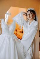 A beautiful bride with a long veil in her room, wearing a robe. Wedding dress on a mannequin. The bride in the morning before the wedding ceremony. photo