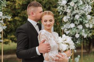 A handsome groom embraces his bride in a lush white dress and smiles in a beautiful outdoor setting. Under the open sky. High quality photo. A newlywed couple poses together on a sunny summer day. photo