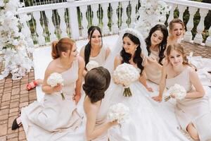 A brunette bride and her bridesmaids in matching cream dresses sit with the bride and rejoice with flowers in their hands near the ceremonial arch. Wedding in nature. photo