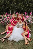 A brunette bride and her bridesmaids in dresses of the same color sit with the bride and rejoice with flowers in their hands near the solemn arch. Wedding in nature. photo