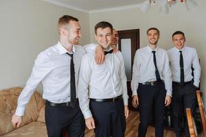 elegante novio riendo y teniendo divertido con padrinos de boda mientras consiguiendo Listo en el Mañana para Boda ceremonia. lujo hombre foto