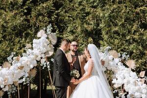 Wedding ceremony in nature. The bride and groom near the flower arch. Master of ceremonies in dark glasses at a wedding during a performance against the background of the bride and groom. photo