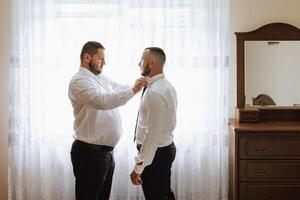 elegante novio riendo y teniendo divertido con padrinos de boda mientras consiguiendo Listo en el Mañana para Boda ceremonia. lujo hombre foto