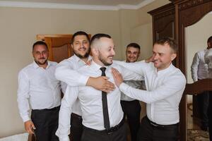 elegante novio riendo y teniendo divertido con padrinos de boda mientras consiguiendo Listo en el Mañana para Boda ceremonia. lujo hombre foto