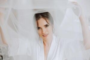 hermosa joven novia participación velo en blanco Boda vestido, retrato de morena novia en hotel habitación, Mañana antes de boda. foto