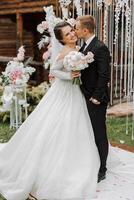 un hermoso novio abraza su novia en un lozano blanco vestir y sonrisas en un hermosa al aire libre ajuste. debajo el abierto cielo. alto calidad foto. un recién casado Pareja poses juntos en un soleado verano día. foto