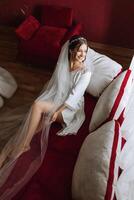 A beautiful bride is sitting in a dressing gown in the morning before the wedding ceremony in a hotel with a modern interior. Incredible hairstyle of the bride. Natural and modern makeup. photo