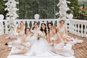 A brunette bride and her bridesmaids in matching cream dresses sit with the bride and rejoice with flowers in their hands near the ceremonial arch. Wedding in nature. photo