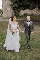 A handsome groom and an elegant bride in a lush white dress are walking in a summer park. Happy bride and groom getting ready for their best day. photo