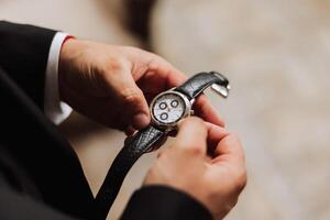 un hombre en un negocio traje sostiene un reloj de pulsera en su manos, un jarra de café es en el mesa en su habitación foto