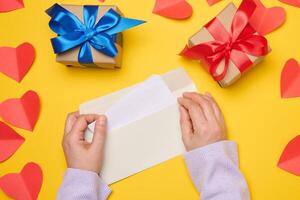 Female hands hold a white rectangular envelope on a yellow background, paper red hearts. V photo