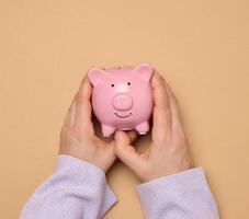 Female hands hold a pink ceramic piggy bank against a brown background photo