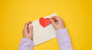 Female hands hold a white rectangular envelope on a yellow background, paper red hearts. photo