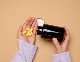 A woman's hand holding a plastic bottle and yellow oval pills on a brown background, top view. photo